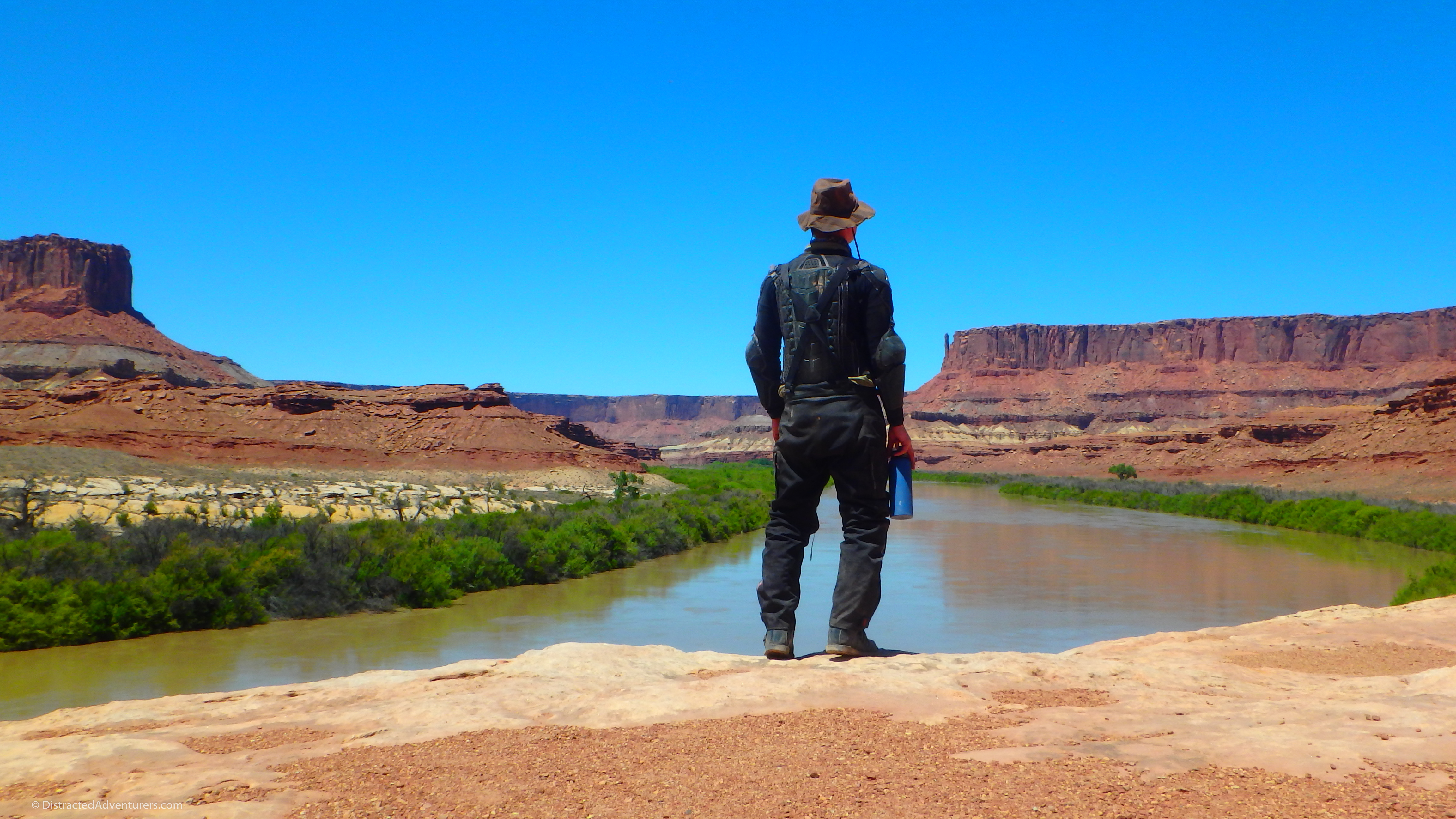 A manly man, Chris, wearing his Indiana Jones style hat and motorcycle body armor stands at the edge of a cliff overlooking a muddy brown river lined by vibrant green trees with large red rock formations in the distance.