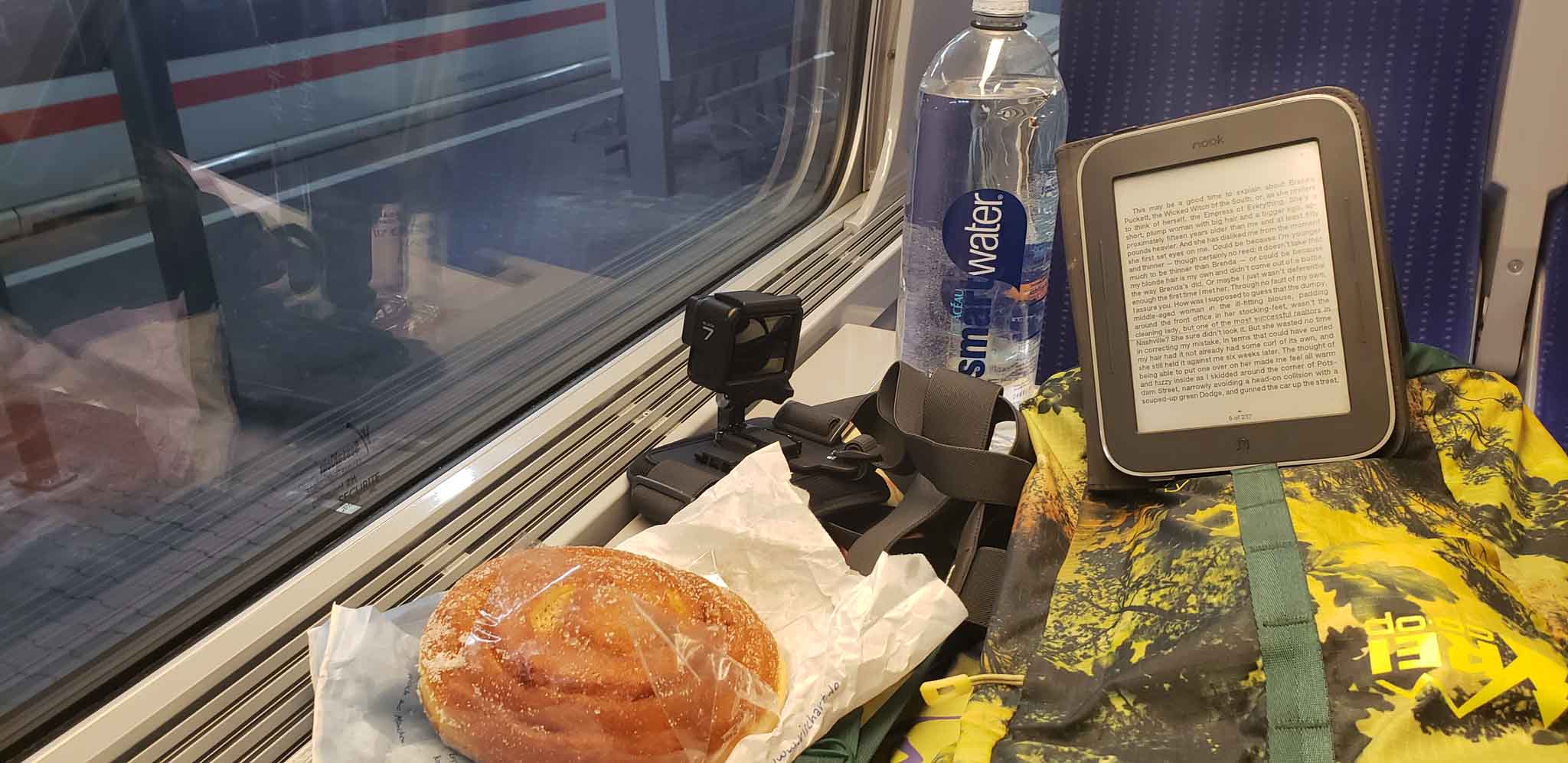 The small train table is covered by a pastry, a Nook e-reader propped up on a green and yellow daybag and a GoPro pointed out the window of the train.