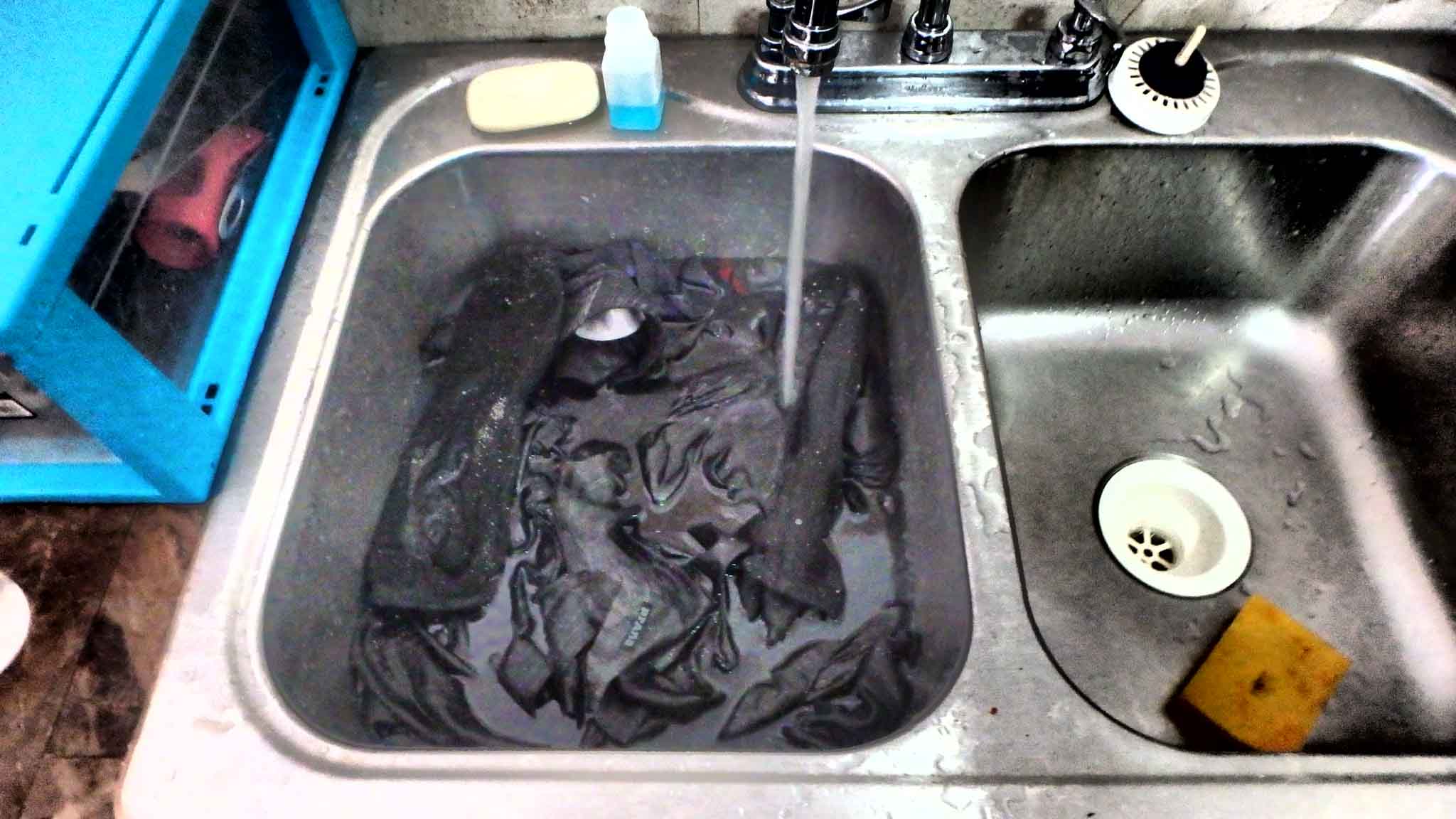 A silver sink holding a bundle of dark clothes is filling with soapy water.